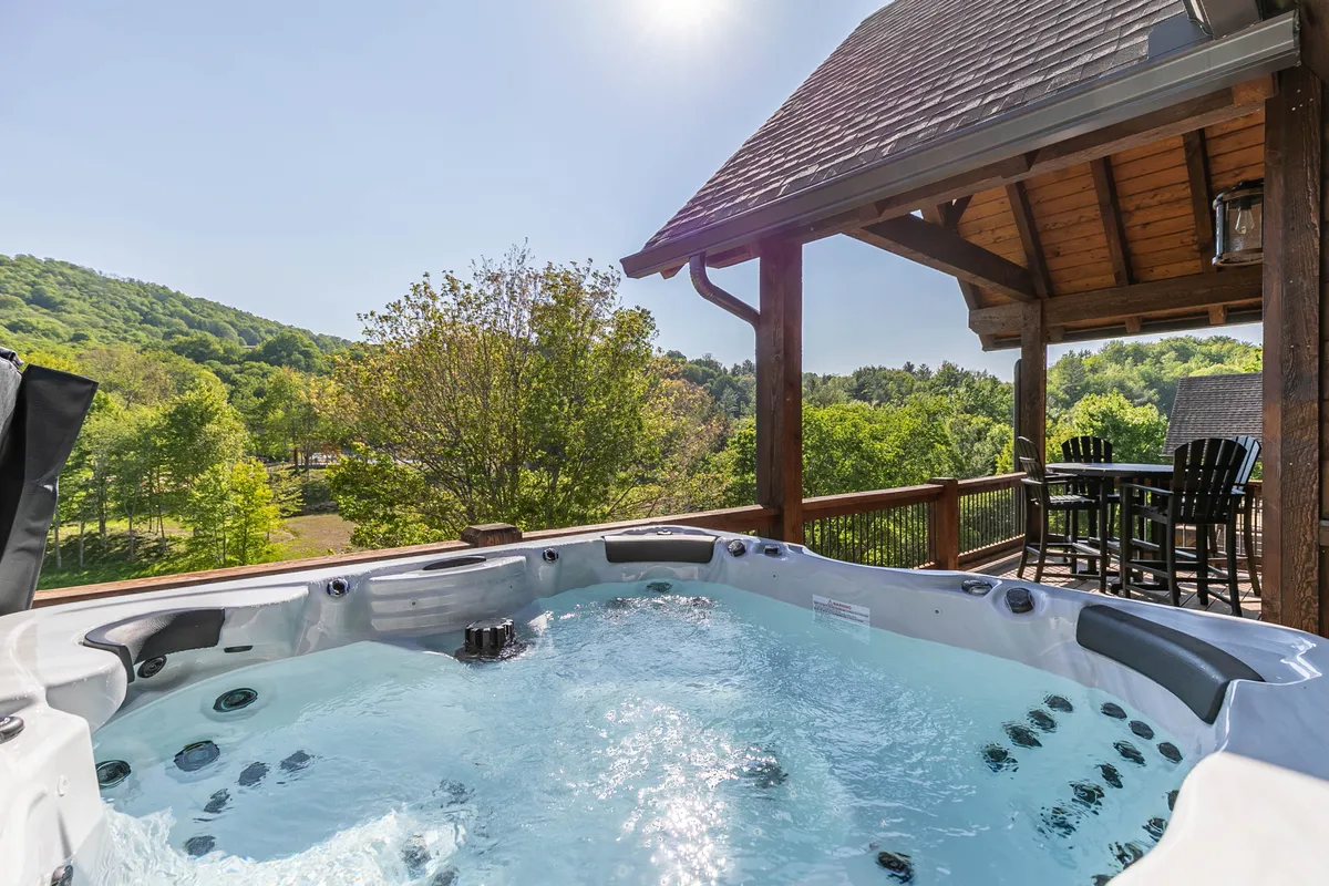 A wide view of the wooden deck featuring the hot tub, ample deck space for entertaining, and the surrounding greenery. The scene evokes serene moments, perfect for enjoying a sunset or gathering with family and friends.