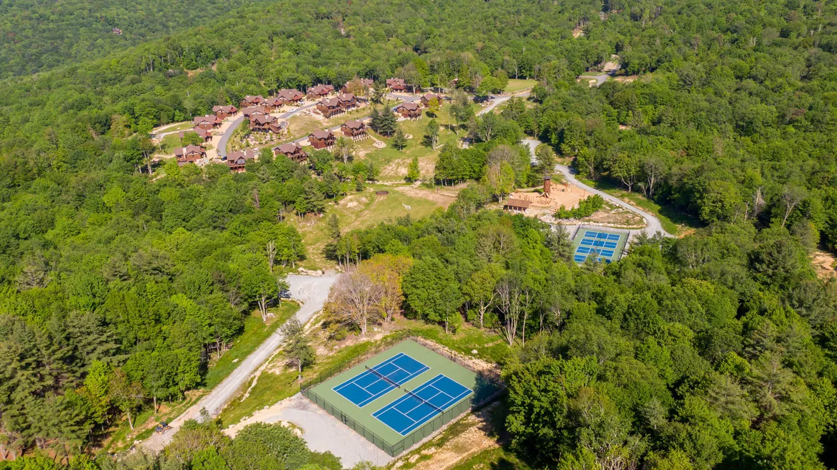 This image showcases a vibrant outdoor scene featuring tennis courts surrounded by wooden cabins nestled under a lush tree canopy. The courts are an inviting space for sports and friendly competition, while the setting hints at potential for community gatherings and outdoor activity.