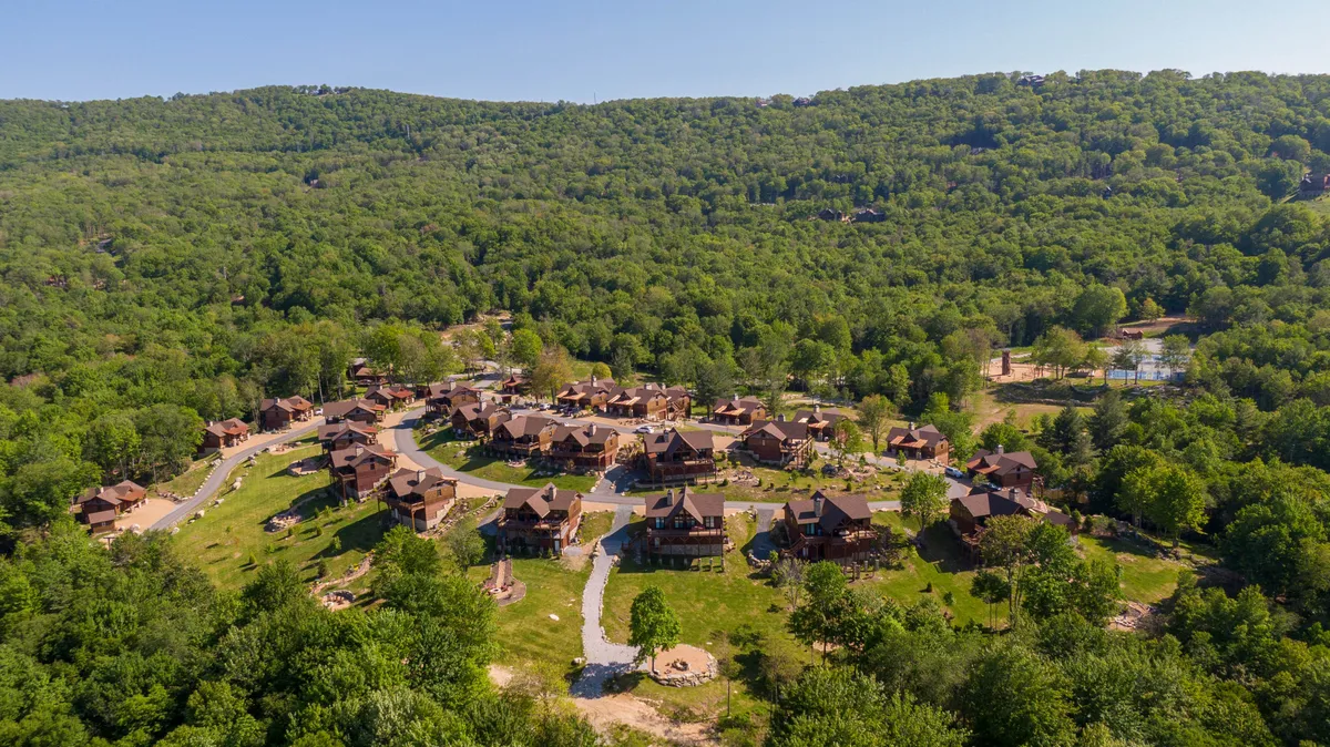 A cluster of wooden cabins nestled among lush, rolling hills and winding pathways. The setting sun casts a golden glow, inviting families to explore the area on foot, enjoy nature walks, or gather for picnics on the hilltop.