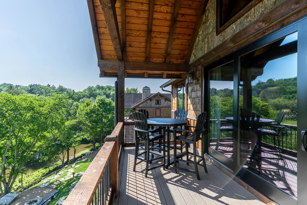 A serene outdoor deck with a wooden balcony railing, an outdoor dining set, lush trees, and a river view. The space is set for al fresco dining, surrounded by natural beauty.