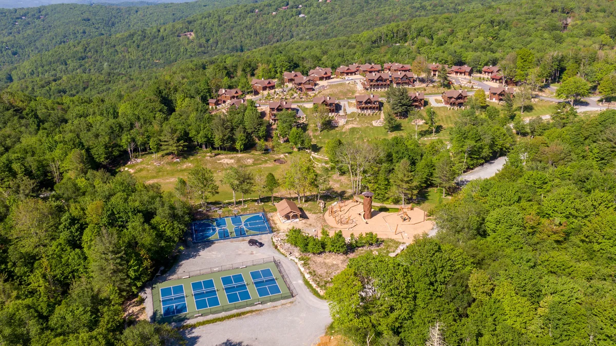 Rows of rustic cabins surrounded by lush greenery, with tennis courts and a playground structure in the foreground. Families and groups are visible enjoying tennis, playing, and preparing for outdoor fun, while the natural backdrop reinforces the mountain location.