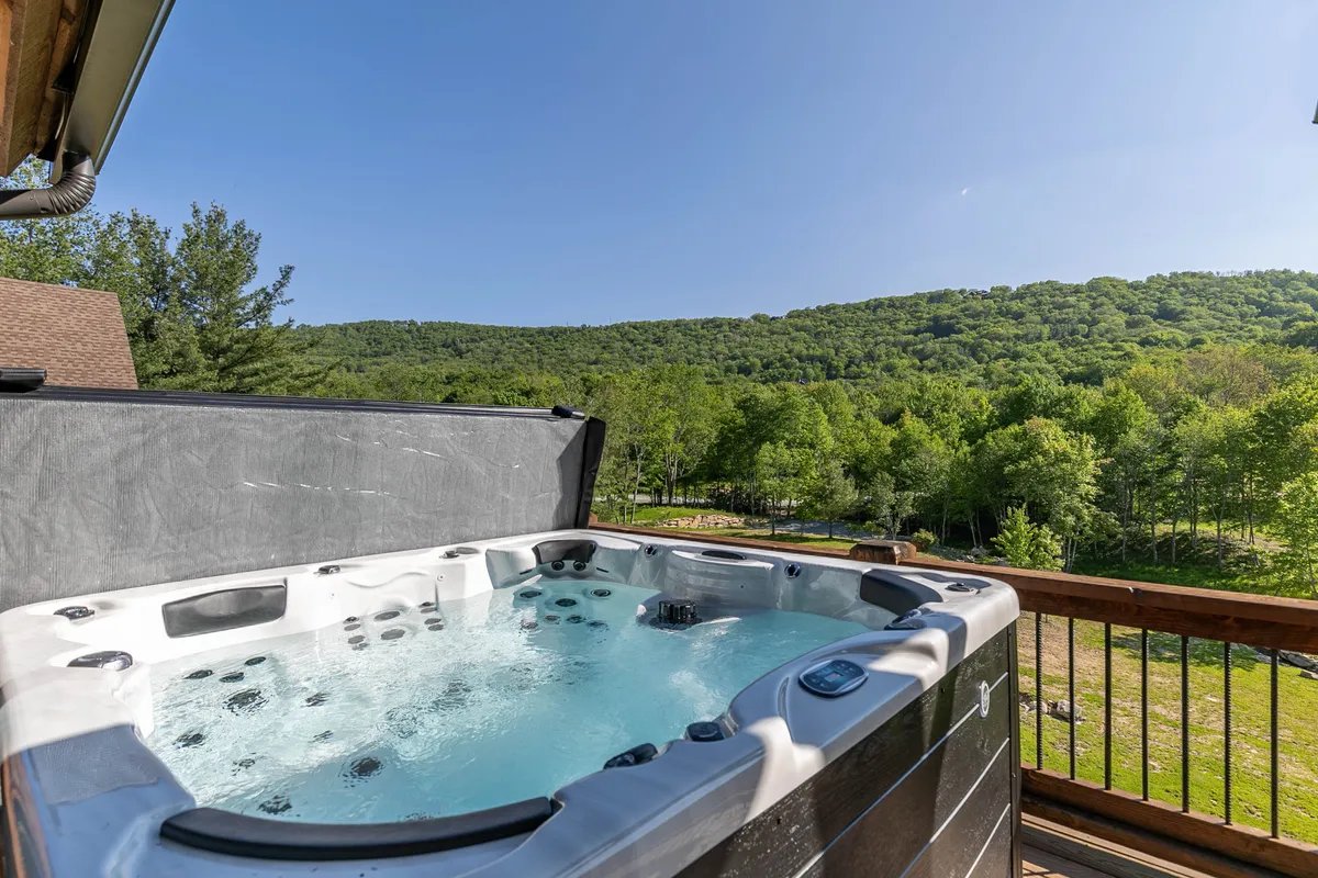 This image showcases a luxurious hot tub positioned on a back deck, surrounded by lush greenery and offering sweeping views of the mountains and valley. The tranquil setting invites relaxation and connection with nature.
