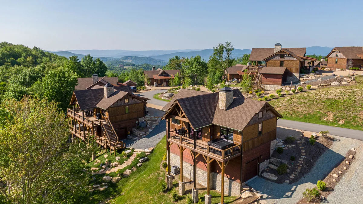The hero image features a charming wooden cabin nestled amid lush greenery, with natural stone pathways leading to the entrance. The cabin is framed by a backdrop of majestic Blue Ridge mountains, with sunlight filtering through the trees and hints of dawn or dusk illuminating the sky. The inviting outdoor space suggests opportunities for evening gatherings, relaxation, and immersion in nature.