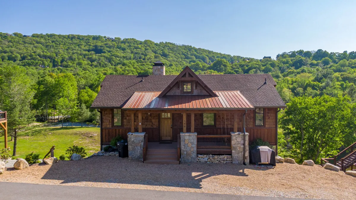 A rustic wooden cabin facade enhanced with stone accents, a welcoming front porch with steps, and surrounded by greenery. The inviting exterior hints at cozy mornings and evenings spent outdoors, enjoying the tranquility of nature.