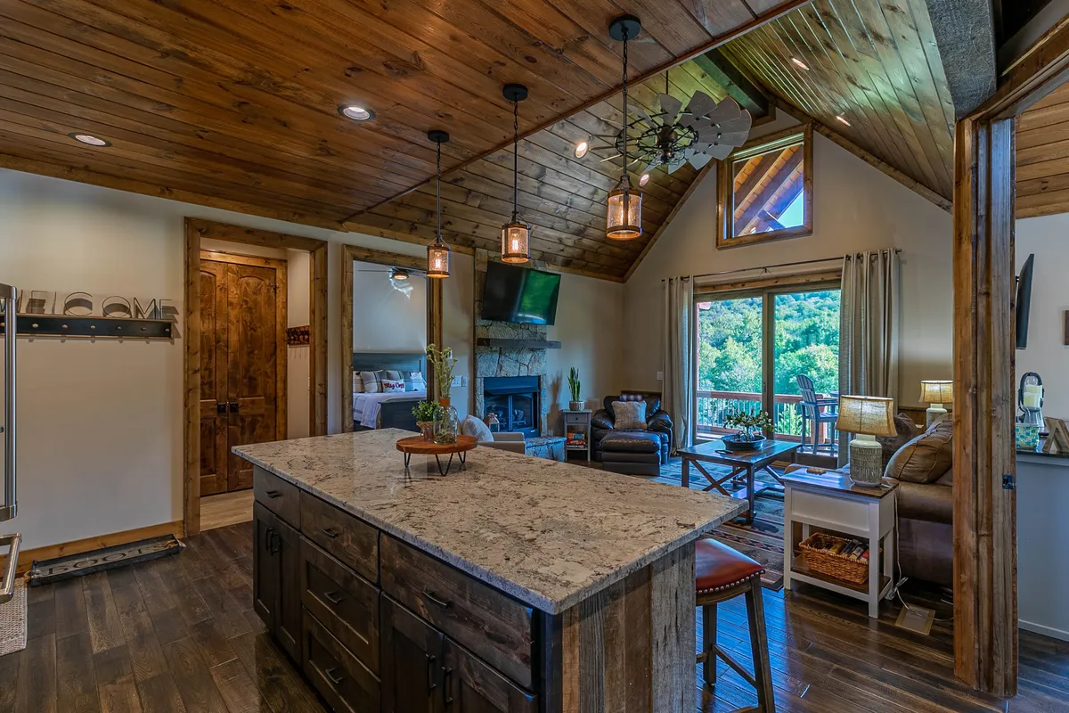 A broad view of an open-concept kitchen and dining space, with an island countertop, a warm fireplace in the background, large windows letting in natural light, and comfortable seating. The setup invites gathering, dining, and conversation.