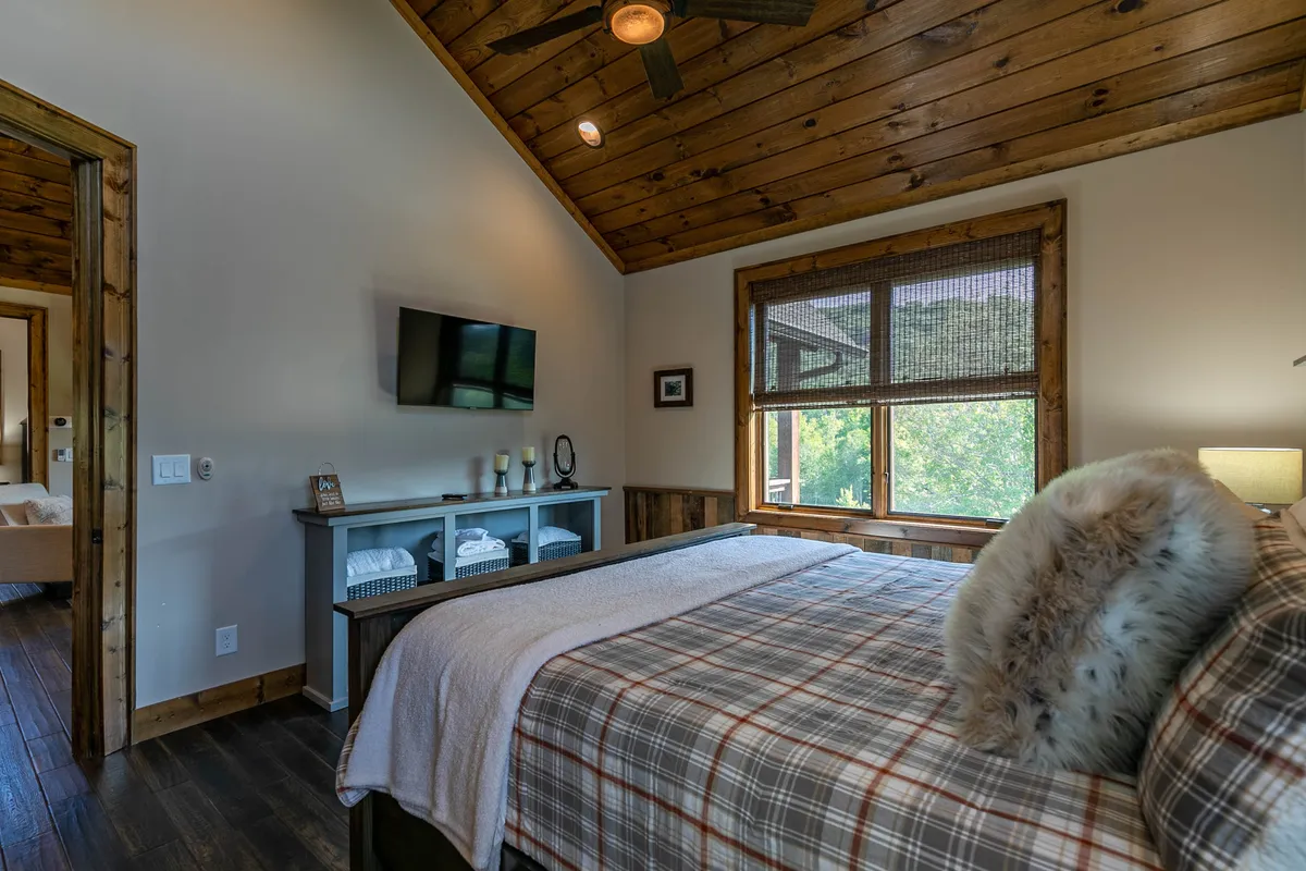 This image showcases a king bedroom with a bed dressed in plaid bedding, a wooden ceiling, a large window with blinds, and a TV mounted on the wall above a decorative console. Natural light streams in, highlighting the inviting and cozy atmosphere of the retreat.