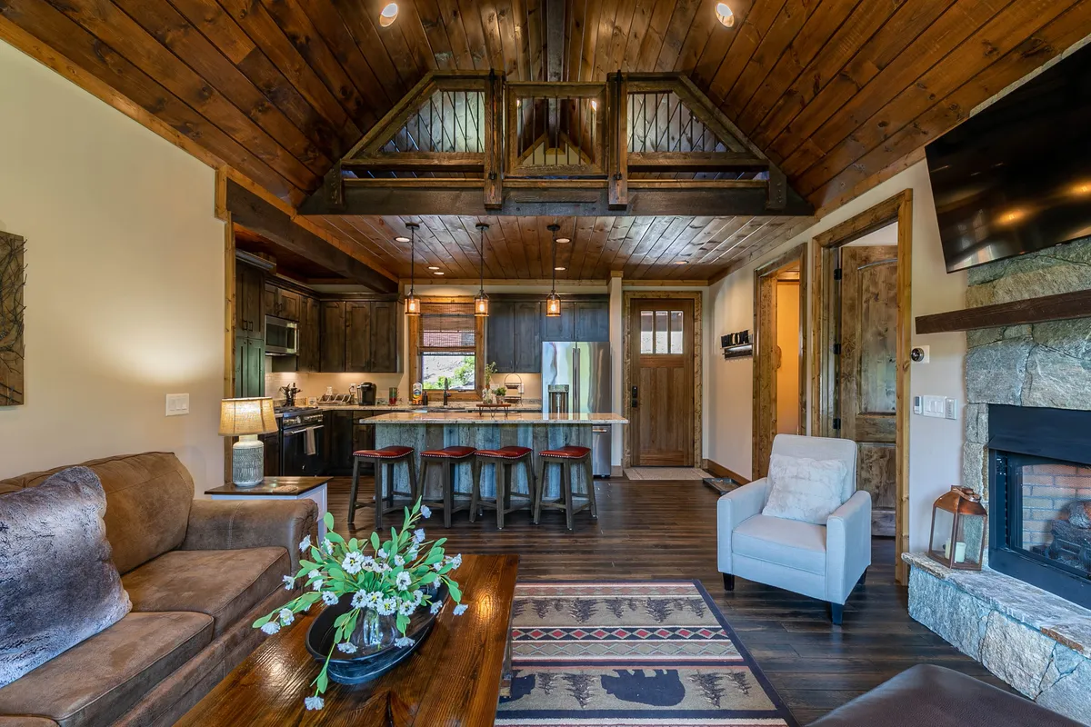 An inviting space with exposed wooden beams, a kitchen island partially visible, a fireplace, plush seating, and decorative touches. The seamless transition between the living area and kitchen hints at an open-plan layout ideal for casual snacking or family meals.